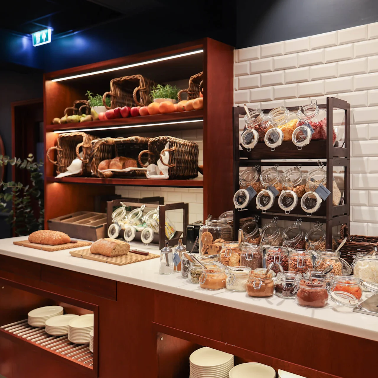 A countertop displays jars of cereals and spreads with loaves of bread. Wicker baskets hold vegetables and fruits on shelves in a warmly-lit kitchen setting.