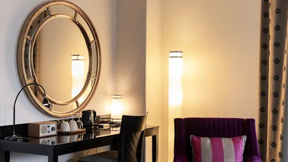Huoneen viihtyisä nurkkaus, jossa on työpöytä, tuoli ja suuri pyöreä peili. Violetti nojatuoli seisoo korkean lampun vieressä.