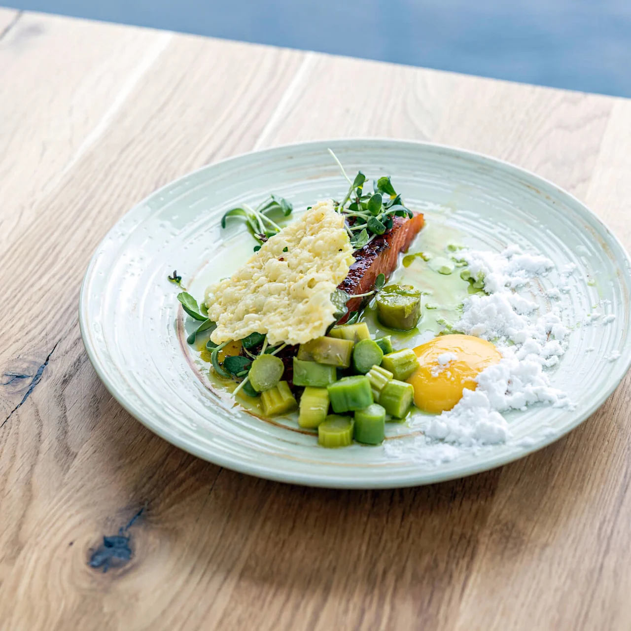 En tallrik med gourmetmat visar ostchips, tärnade grönsaker, äggula och mikrogrönt, elegant arrangerade mot ett träd bordsunderlag vid vattnet.