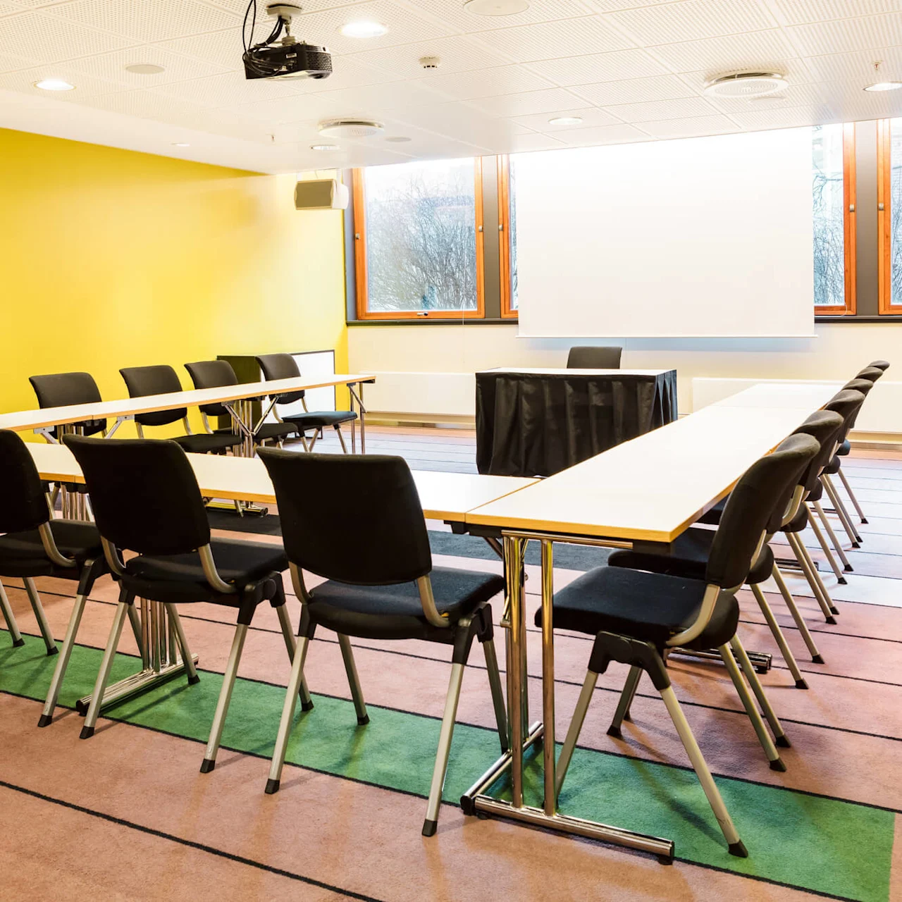 Conference room with black chairs arranged around U-shaped tables. A desk with a draped cover and a projector screen are against a yellow wall, large windows in background.