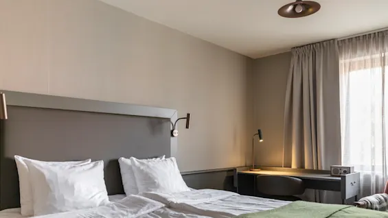 Kutsuva hotellihuone, jossa on siististi pedattu sänky, hyvin valaistu työpöytäalue ja pehmeä valo suodattuu ikkunan läpi.