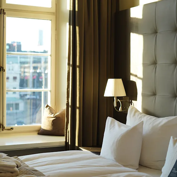 A bright hotel room with a neatly made bed, a window overlooking a city, and warm sunlight streaming in.