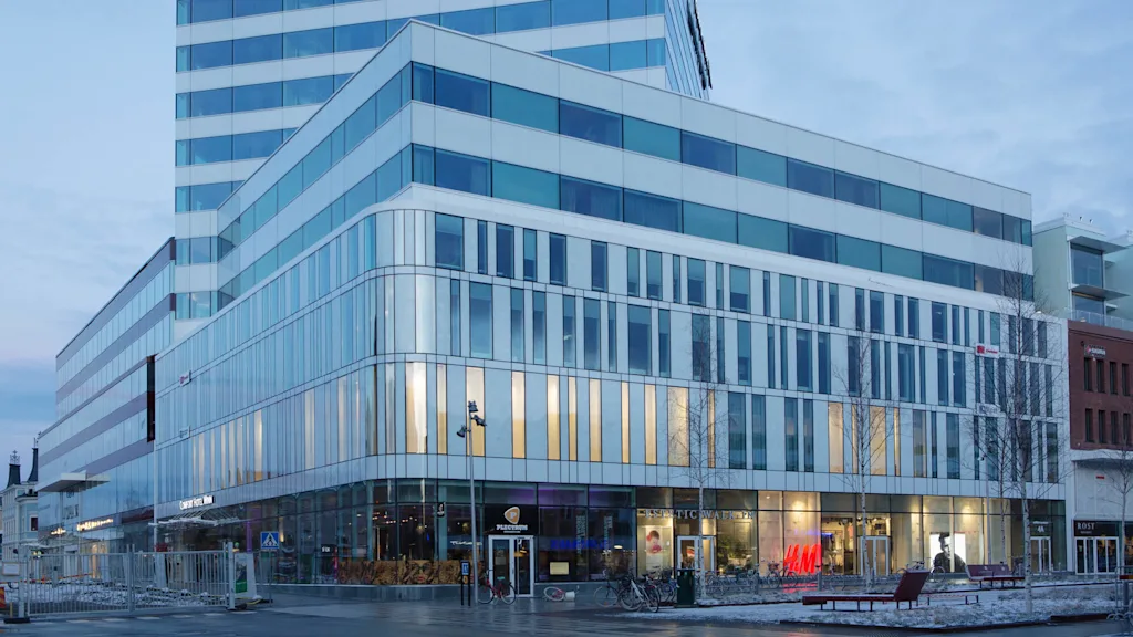 Nykyajan monikerroksinen hotellirakennus, lasijulkisivu, valaistu sisätila, iltahämärän taivas.