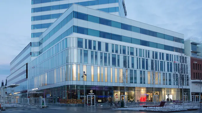 Nykyajan monikerroksinen hotellirakennus, lasijulkisivu, valaistu sisätila, iltahämärän taivas.