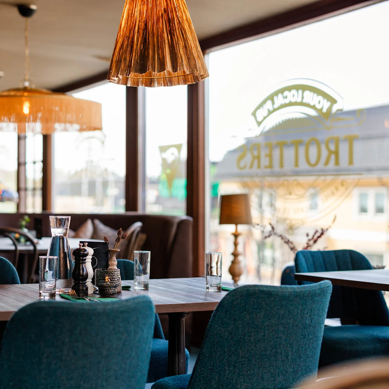 Matbord dukat för servering med glas och kryddor, placerat i ett mysigt kafé med stilfull belysning. Stora fönster visar texten baklänges: "TROTTERS" och "YOUR LOCAL PUB."