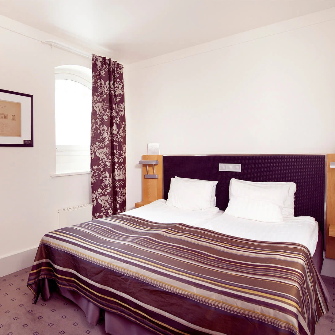 A neatly made bed in a tidy hotel room with a striped comforter, matching curtains, and a framed artwork on the wall.