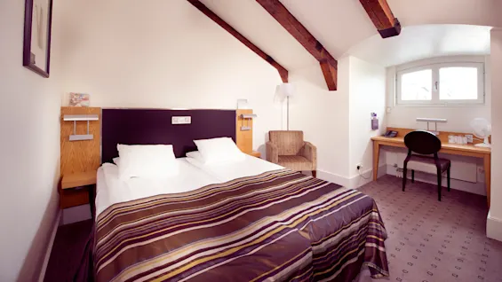 A neatly arranged hotel room with a double bed, exposed wooden beams, and a work desk in a cozy setting.