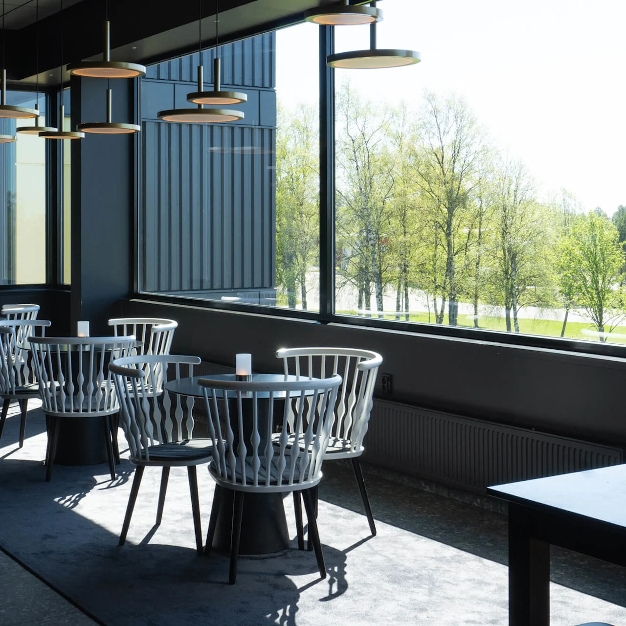 Bord med svarta toppar och vita stolar under runda taklampor på ett modernt inomhuscafé; stora fönster visar en vy av gröna träd utanför.