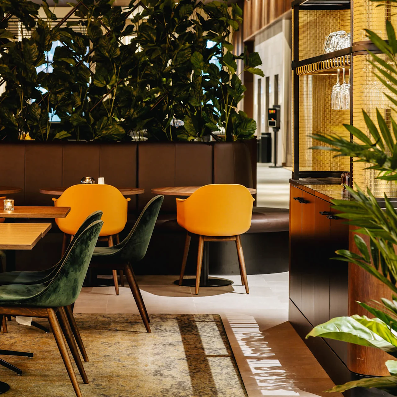 Borde med grønne og orange stole er arrangeret i et hyggeligt café-miljø, omgivet af frodige indendørsplanter. Blød belysning fremhæver den varme, indbydende atmosfære.