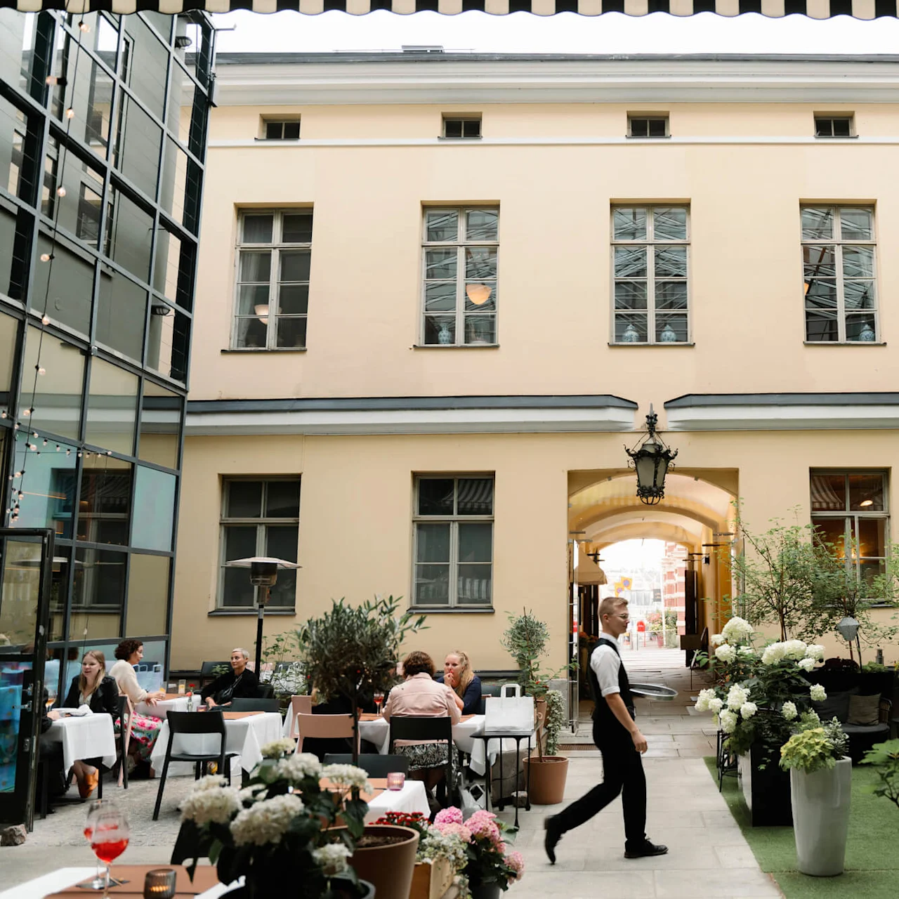 Outdoor restaurant patrons sit at tables with plants and string lights around. A waiter walks past in a courtyard surrounded by buildings and greenery.
