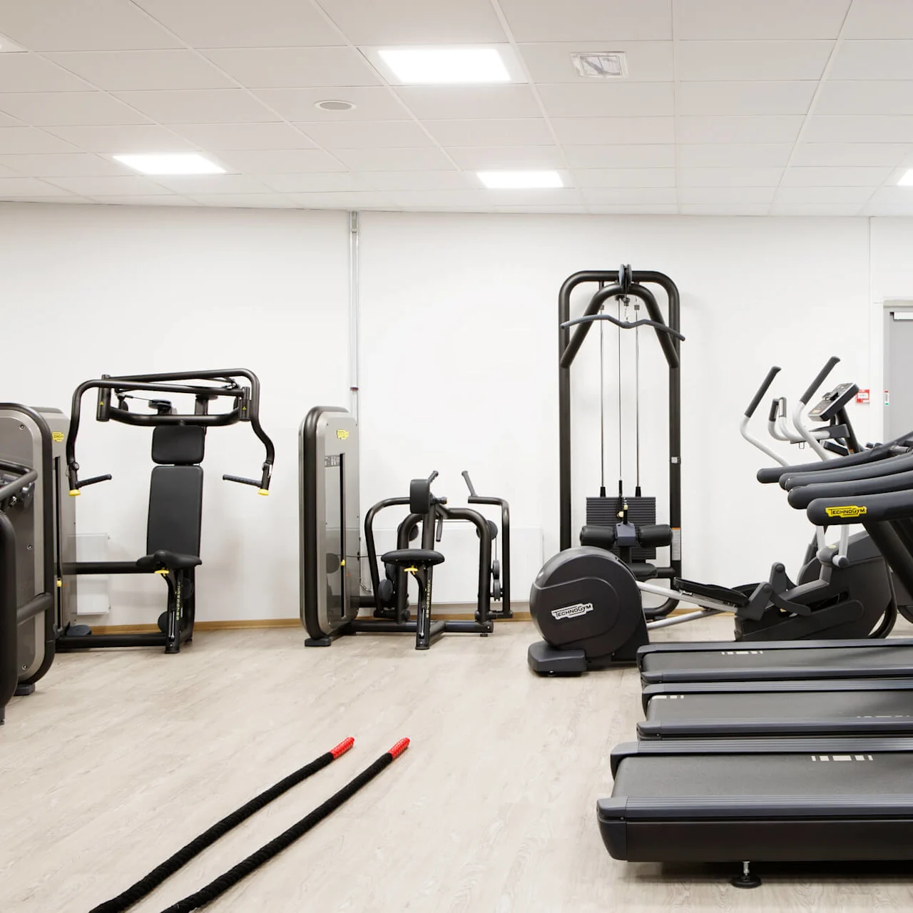 Treadmills line the right side of a clean, orderly gym. Exercise machines are positioned on the left. The room is bright with white walls and ceiling lights.
