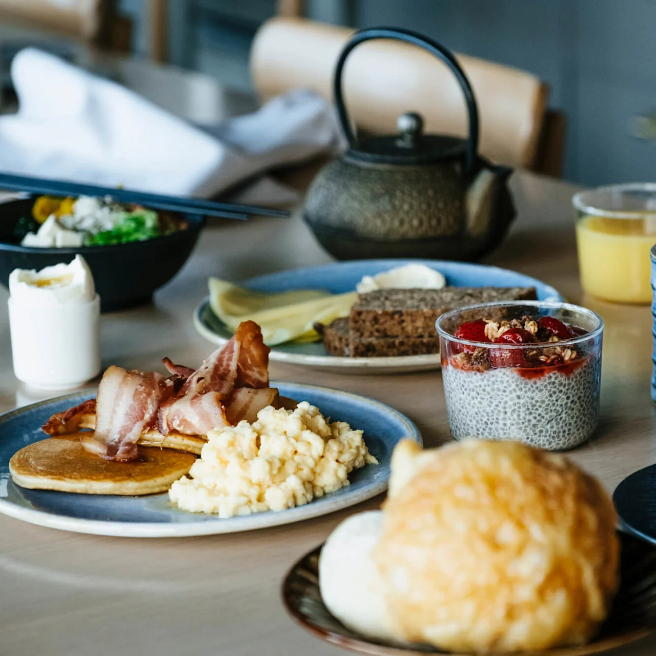 Breakfast dishes including scrambled eggs, bacon, pancakes, bread with toppings, chia pudding, and drinks are set on a table with a teapot in a cozy kitchen setting.