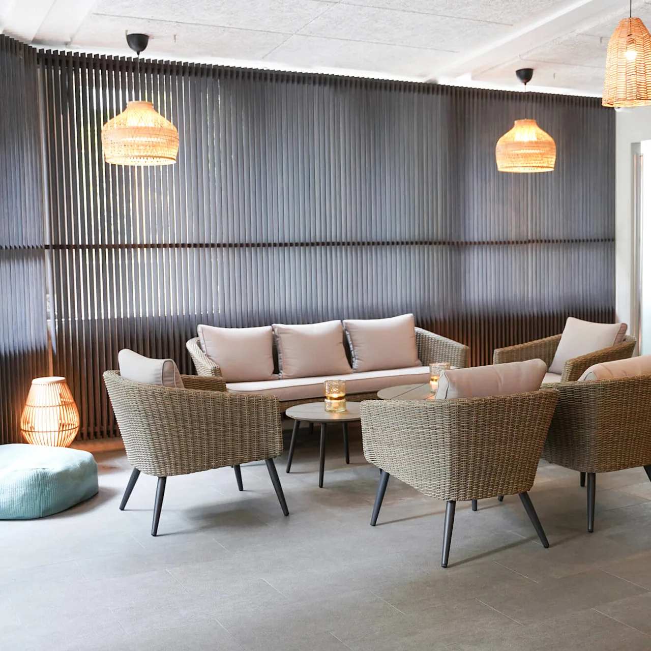 Wicker chairs and sofa with cushions surround a round table in a lounge with modern rattan lights and a wood-paneled wall.