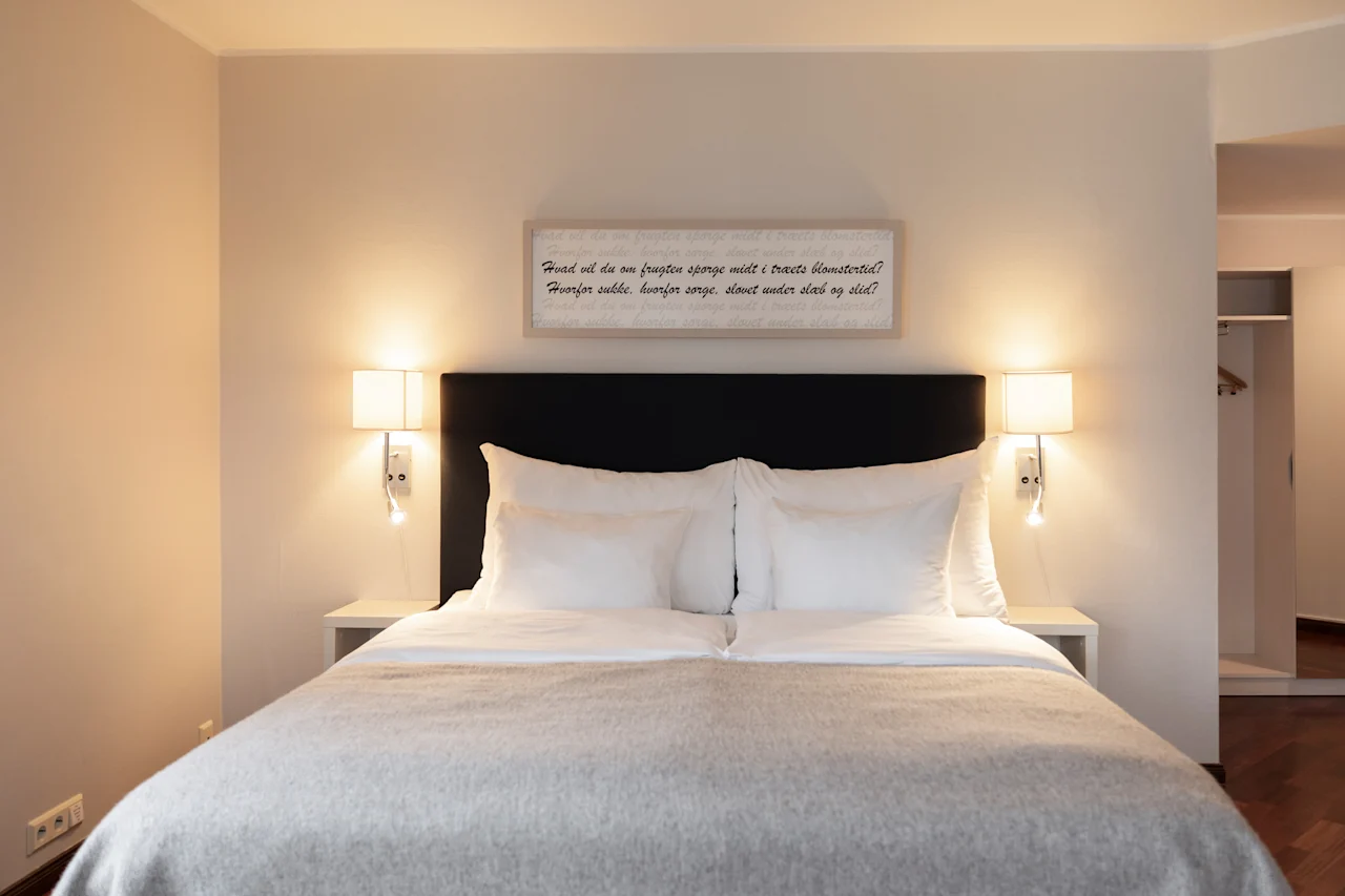 A serene hotel room featuring a comfortable bed with crisp white linens, soft lighting from wall lamps, and a framed quote above.
