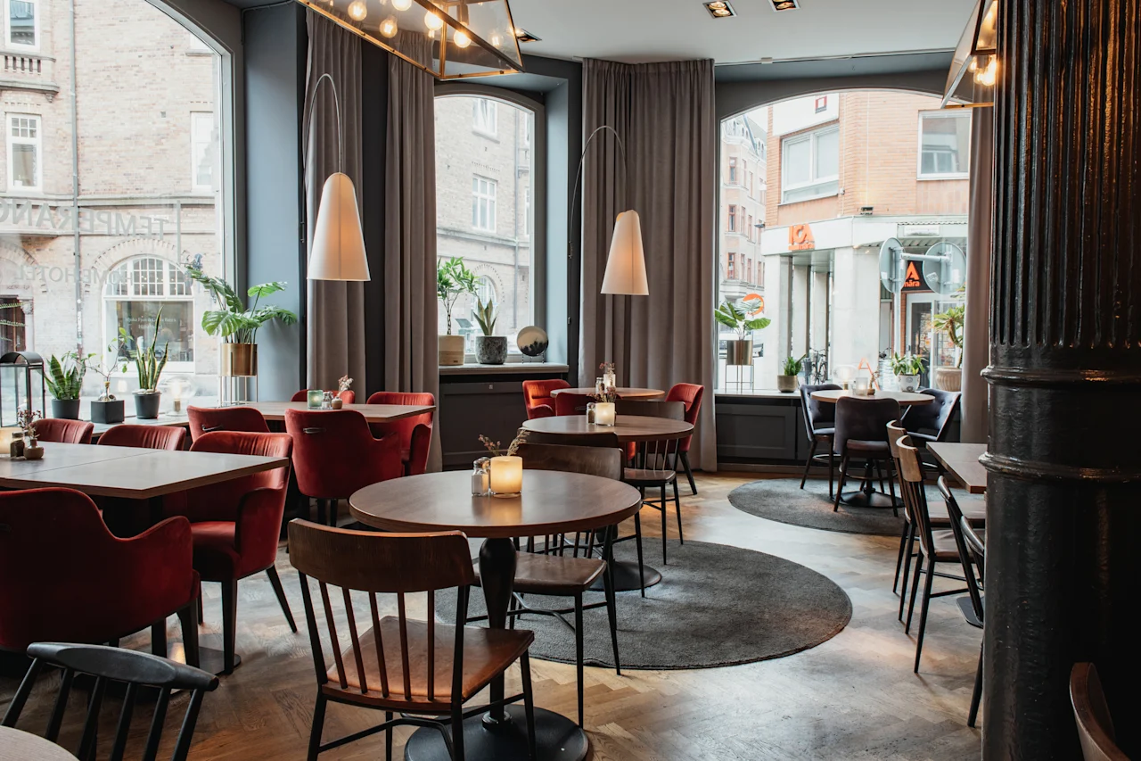 Et stilfullt restaurantinteriør med ulike sitteplasser, levende lys på bordene og store vinduer mot en bygata. HOTEL TEMPEL.