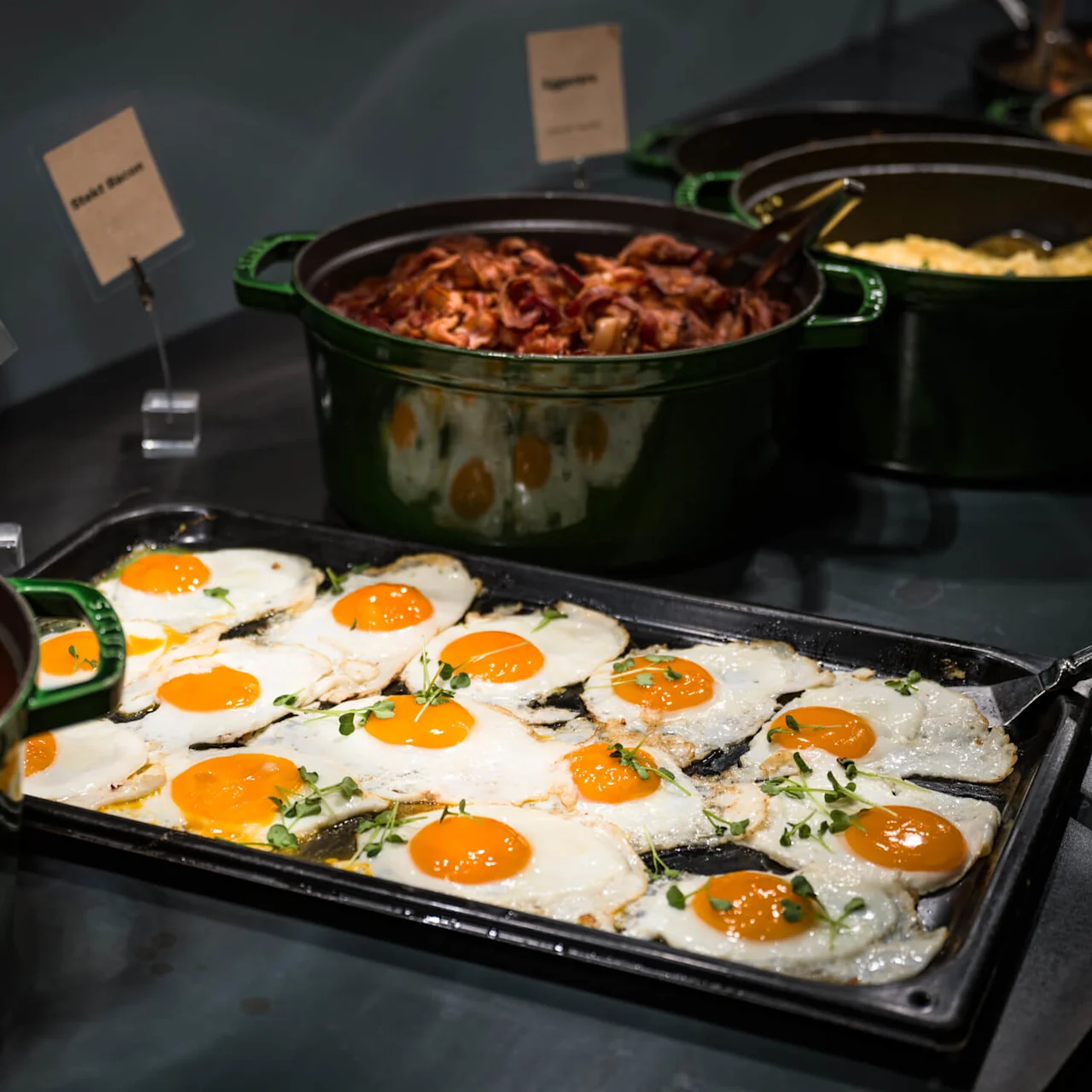 Stekte egg ligger på en stekeplate med urter, omringet av gryter med bakte bønner og bacon i en buffet. Skilt leser "Speilegg" og "Stekt Bacon".