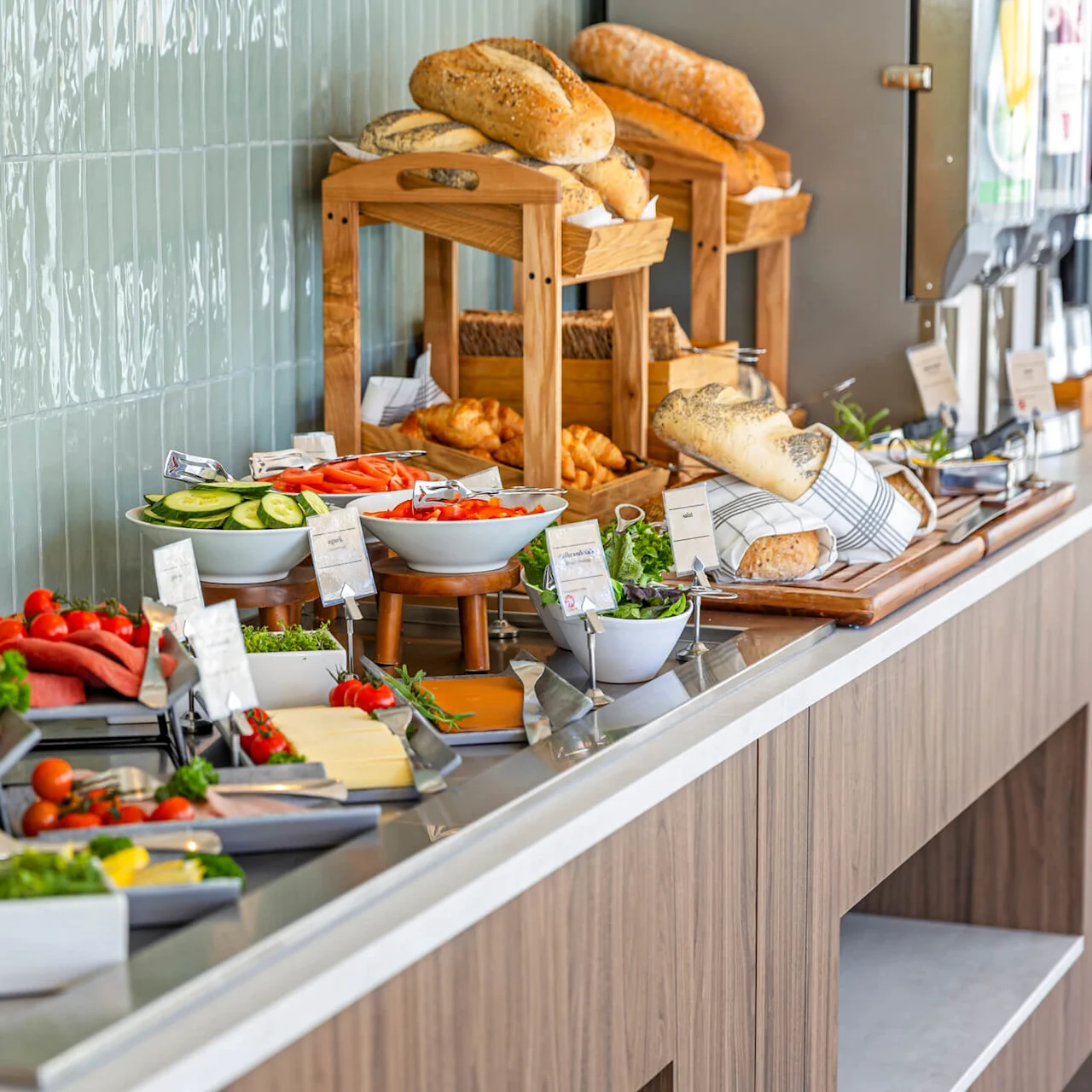 A breakfast buffet with assorted bread, meats, cheeses, and vegetables, displayed in an orderly fashion with labels in front of a tiled wall.