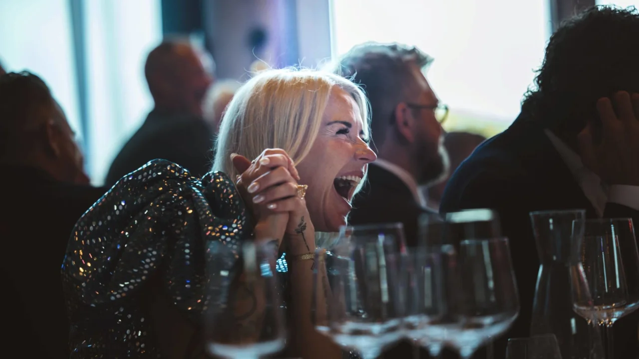 A woman laughing heartily amidst a social gathering, with wine glasses in the foreground.