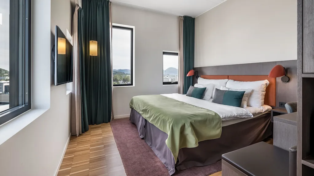 Siiististi pedattu sänky vihreällä peitteellä sijaitsee modernissa hotellihuoneessa, jossa on suuret ikkunat, tummat verhot, puulattia ja seinälle kiinnitetty televisio.