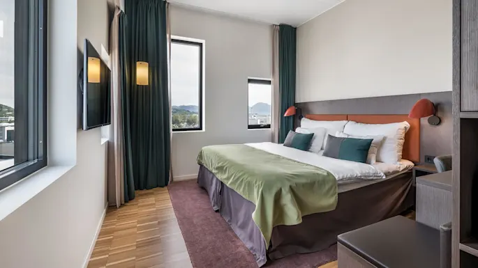 A neatly made bed with green cover stands in a modern hotel room with large windows, dark curtains, wooden floor, and a wall-mounted TV.