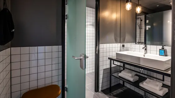 Et bad med hvitkledde fliser og en moderne vask på en svart metallramme. Håndklær er pent stablet nedenfor. En frostet glassdør er delvis åpen.