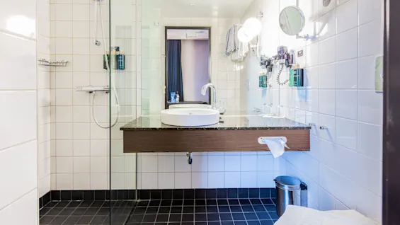 A modern bathroom features a white sink on a wooden counter with a mirror and bright lighting. A glass shower enclosure is to the left, and towels are hanging nearby.