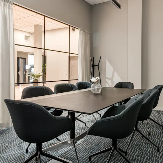 A conference table with six dark chairs surrounds a small vase of flowers. The space features large windows with sheer curtains, and the adjacent hallway is visible.