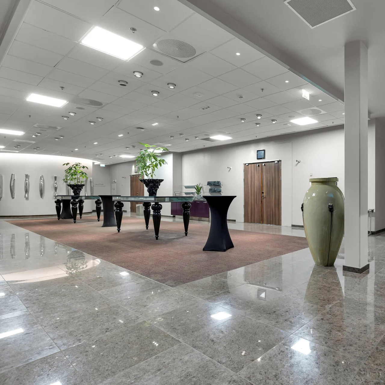 En rummelig gang med skinnende flisegulv har et stort, dekorativt bord med grønne planter. Vægge pyntet med moderne kunst og en trappe med glasgelændere fuldender det elegante rum.