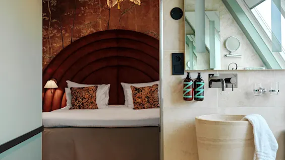 A luxurious bedroom features an arched headboard bed with patterned pillows. Adjacent is a modern bathroom with a stone sink and stylish toiletries against a backdrop of large windows.