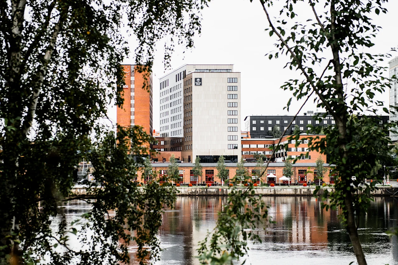 Höga byggnader står på andra sidan en flod, sedda genom löviga trädgrenar, vilket skapar ett inramat stadslandskap.