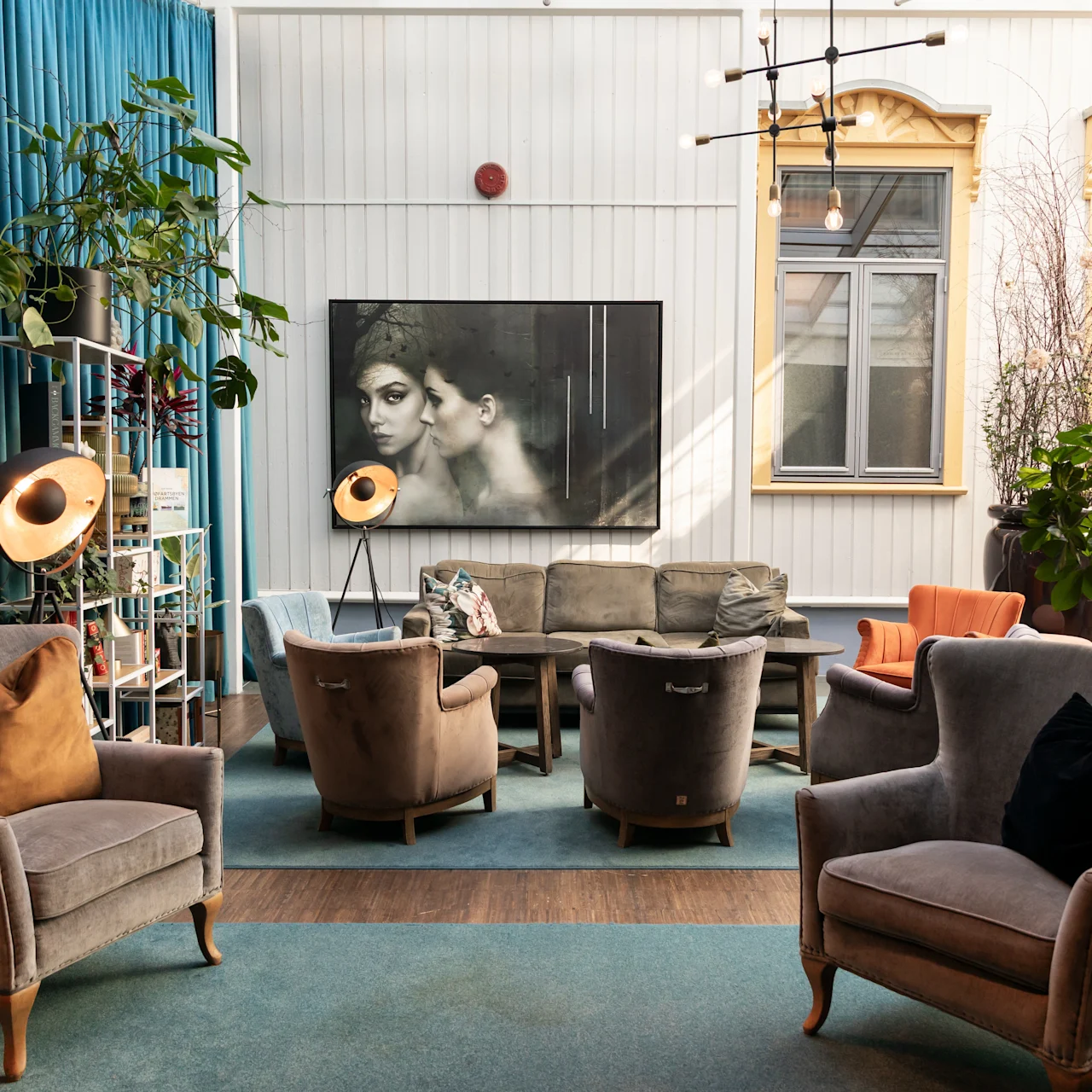 Grå lænestole omkranser et sofabord, mens et stort kunstværk pryder væggen. Frodige planter og industrielle lamper skaber en hyggelig, kunstnerisk lounge-atmosfære med blå gardiner og træaccenter.