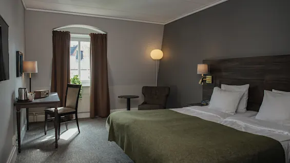 Ett prydligt arrangerat hotellrum med en dubbelsäng med vita lakan och en grön filt. Ett skrivbord och stol står vid ett fönster med fördragna gardiner i en mjukt upplyst, gråmålad miljö.
