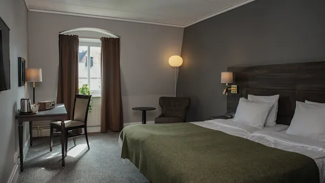 A neatly arranged hotel room features a double bed with white linens and a green blanket. A desk and chair sit by a window with drawn curtains inside softly lit, gray-walled surroundings.