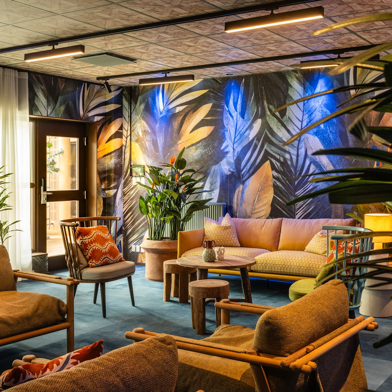 Comfortable seating area with wooden chairs and sofas, featuring tropical-themed wallpaper and surrounded by lush green plants, creating a cozy and vibrant atmosphere.