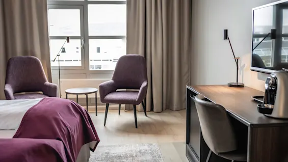 Et hotelværelse med to lilla stole foran et lille rundt bord. I nærheden en seng med lilla tæppe og et skrivebord med lampe og kaffemaskine, under et vægmonteret TV.
