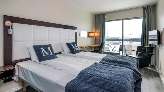 Ett hotellrum med två enkelsängar, varje med kuddar med "M" på, har utsikt över stadsmiljön genom stora fönster. Ett skrivbord, en stol och en fåtölj förbättrar dess funktionalitet.
