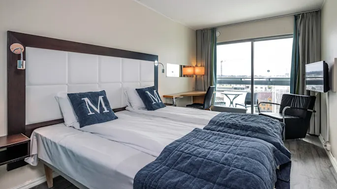 A hotel room with twin beds, each having pillows with "M" on them, overlooks a cityscape through large windows. A desk, chair, and armchair enhance its functionality.