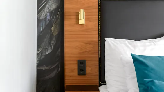 A bedside with a wood panel, gold light switch, and power outlets against an abstract wallpaper, with a pillow and teal cushion.