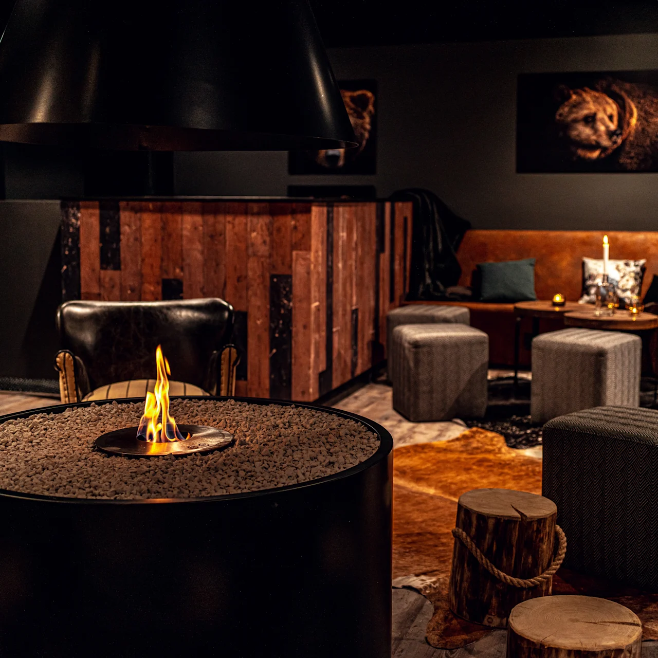 A circular fire pit sits in a cozy lounge with leather chairs and a brown sofa. Wooden stools and gray ottomans surround it, with animal paintings on dark walls.