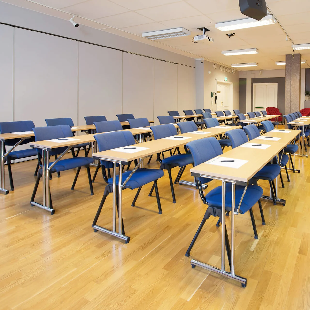 Blå stolar och bord med papper i ett tomt klassrum, parallellt med en presentationsduk framtill.