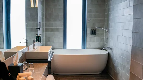 Modern bathroom features a freestanding bathtub beneath two tall windows, with a sleek sink and counter on the left. Grey tiles cover the walls and floor for a minimalist design.