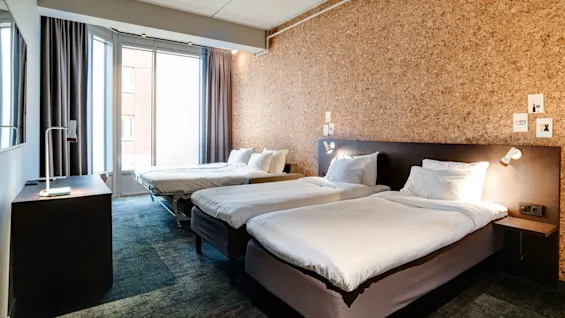 A hotel room with three neatly made beds, cork wall with small pictures, large windows with curtains, and a desk with a lamp. A carpet covers the floor.