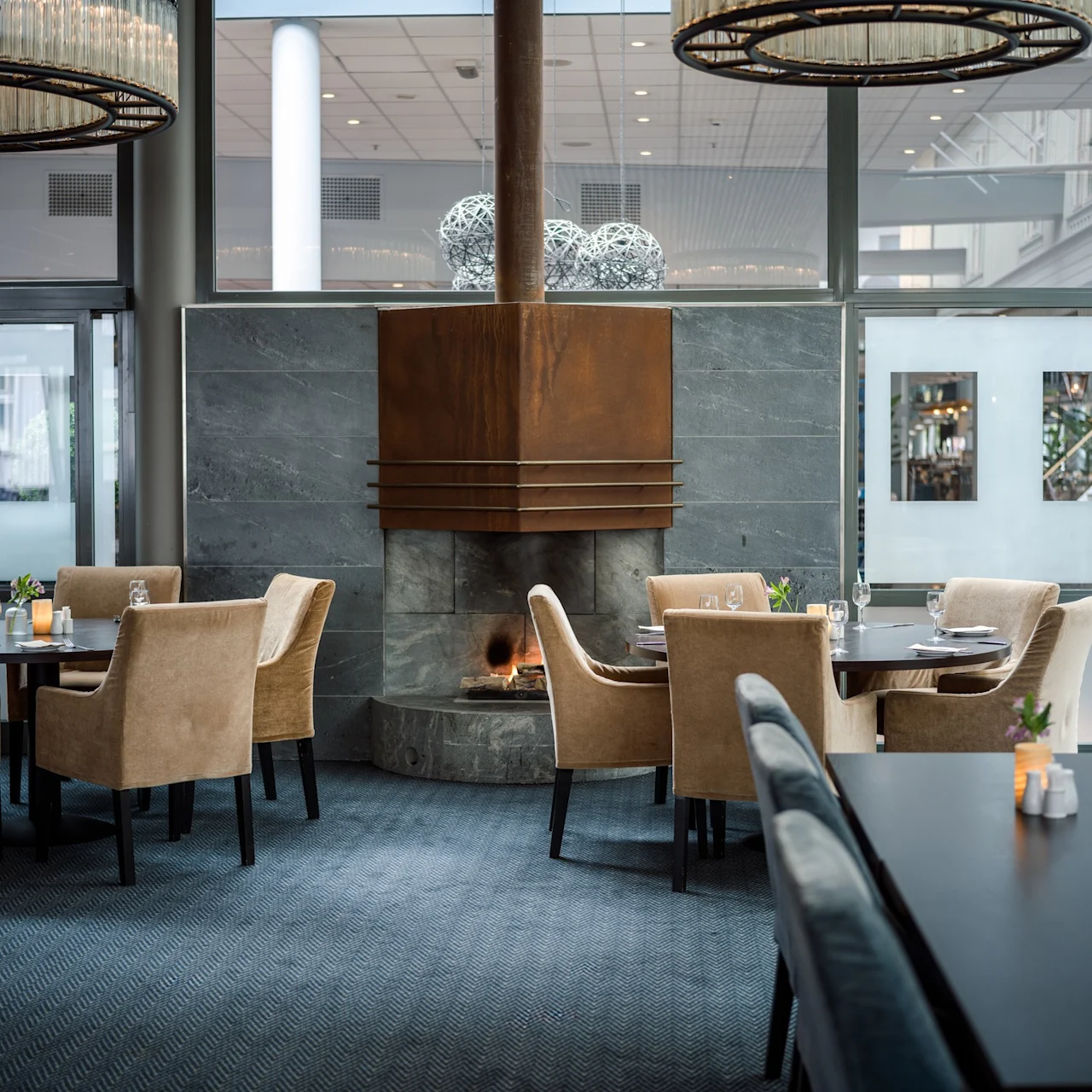 Matsalsstolar omger bord dukade med glas och ljus, placerade nära en tänd eldstad i en modern restaurang med stora fönster och dekorativ belysning.