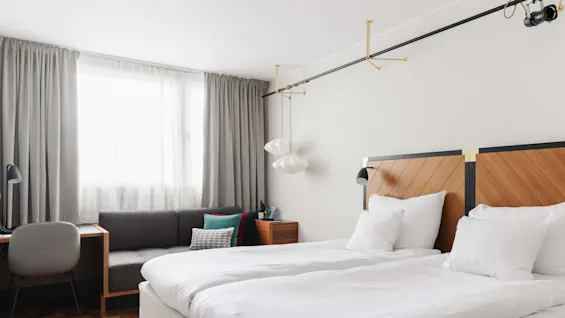Modernt sovrum med en snyggt bäddad säng, stilfulla ljusarmaturer och en sittplats med en grå soffa, i ett rent och ljust utrymme.