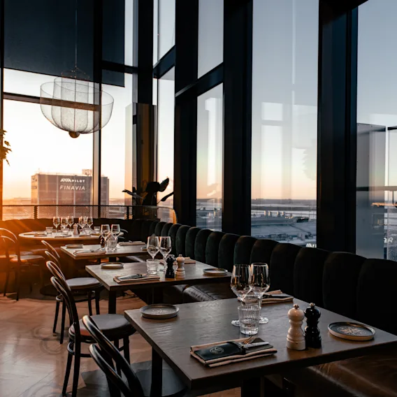 Ravintola NÒR Clarion Hotel Helsinki Airportissa, katetut pöydät, kylpee lämpimässä auringonlaskun valossa suurista ikkunoista.