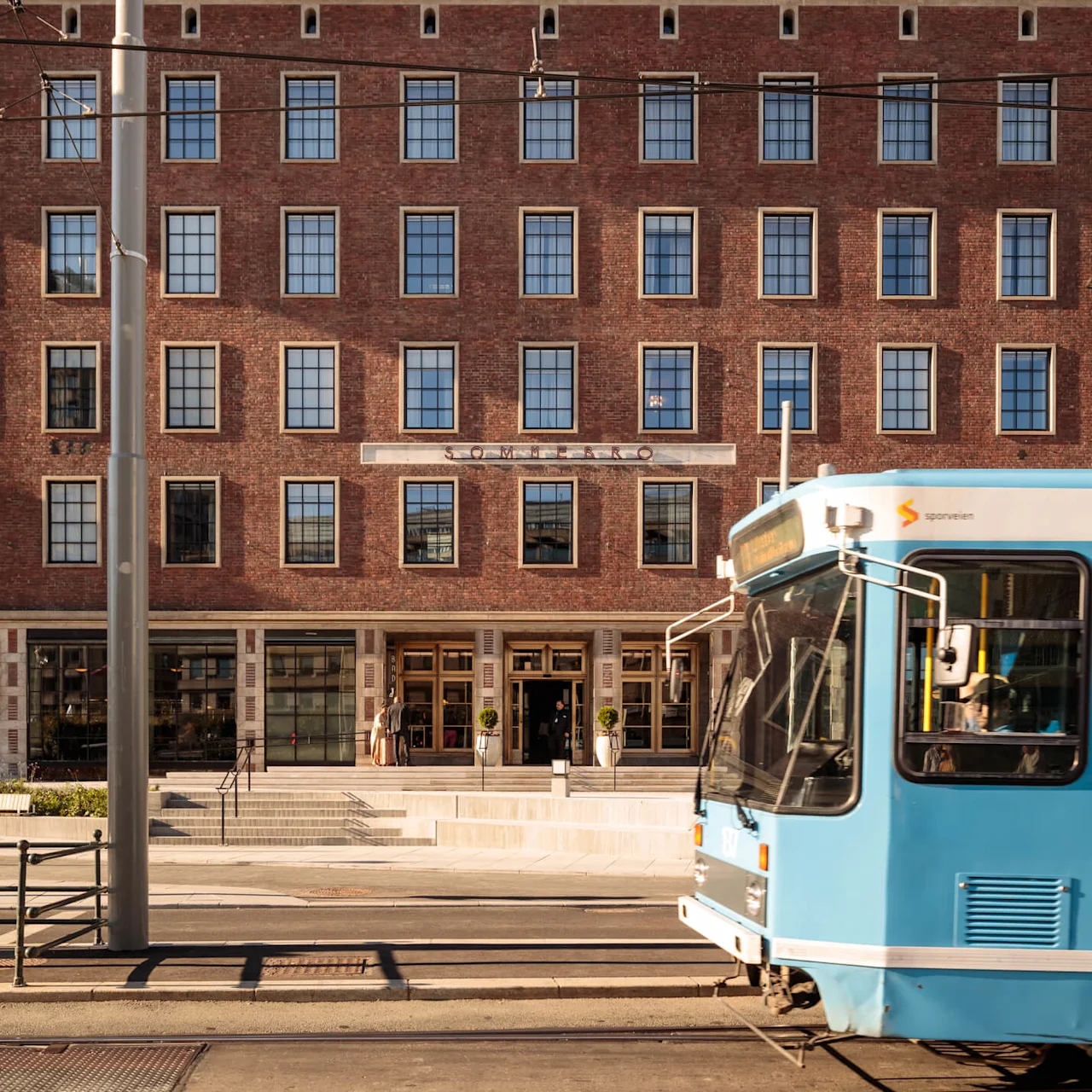En sporvogn passerer foran en murstensbygning med store vinduer og "SOMMERRO"-skiltet over indgangen, i et solrigt bymiljø.