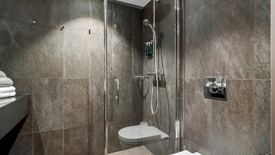 En glasslukket bruser i et moderne badeværelse har et regnbruserhoved og håndholdt dyse, med mørke flisevægge og en indbygget hylde med toiletartikler. En stabel håndklæder hviler på disken.