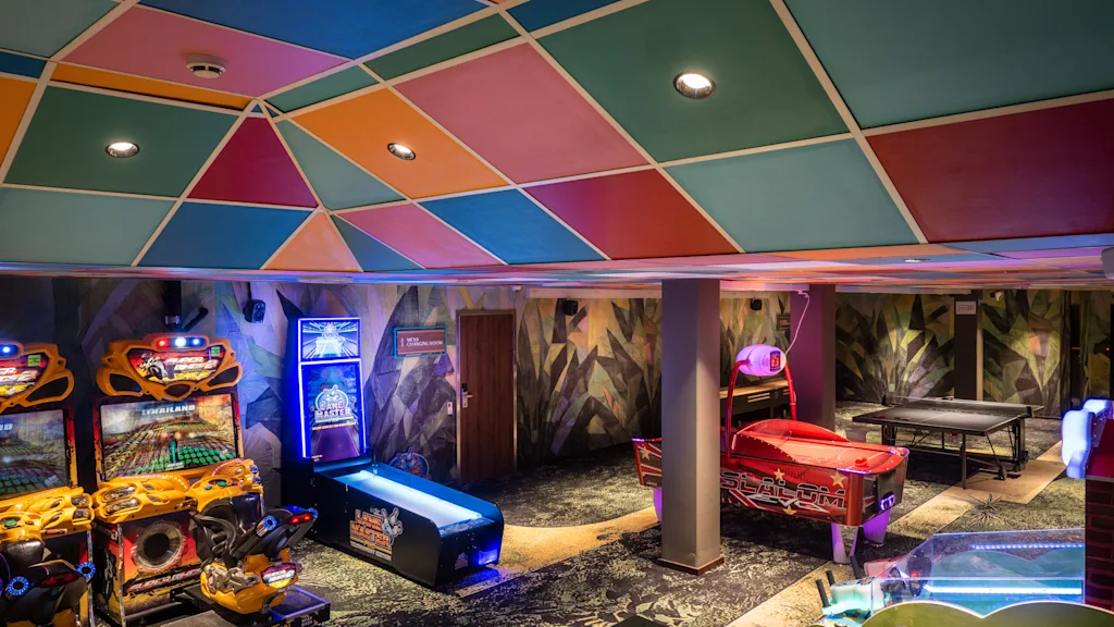 Arcade games stand in a colorful room, including racing games, air hockey, and pinball machines, under a vibrant, patterned ceiling. Neon lights illuminate the space, creating an exciting gaming environment.