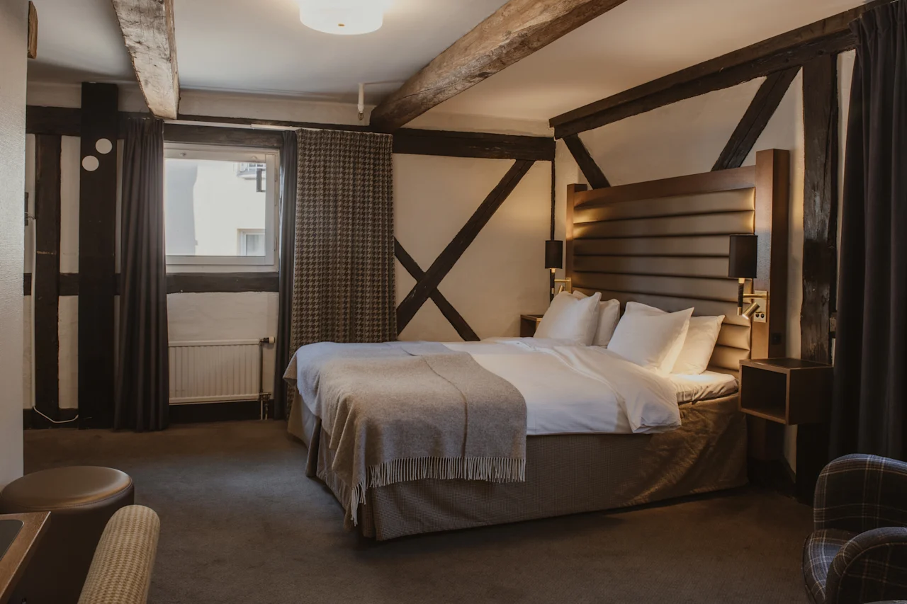 Et hyggeligt hotelværelse med en stor seng, synlige træbjælker og blød belysning, perfekt til et afslappende ophold.