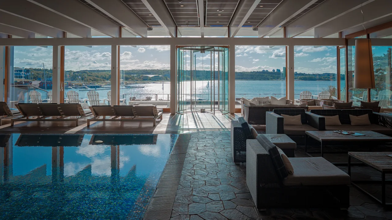 Indendørs poolområde med liggestole og siddepladser, med panoramaudsigt over en solrig sø med både og en fjern kystlinje.